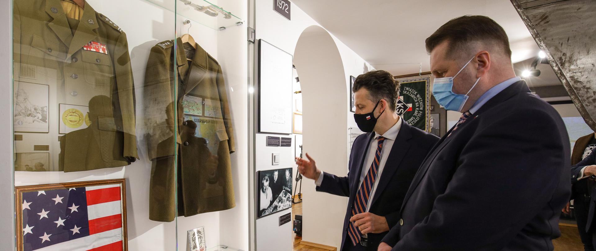 Minister Przemysław Czarnek i dyrektor muzeum stoją przed szklaną gablotą. W gablocie umieszczone są dwa mundury, dwie czapki żołnierskie, buty, aparat oraz inne rekwizyty. 
