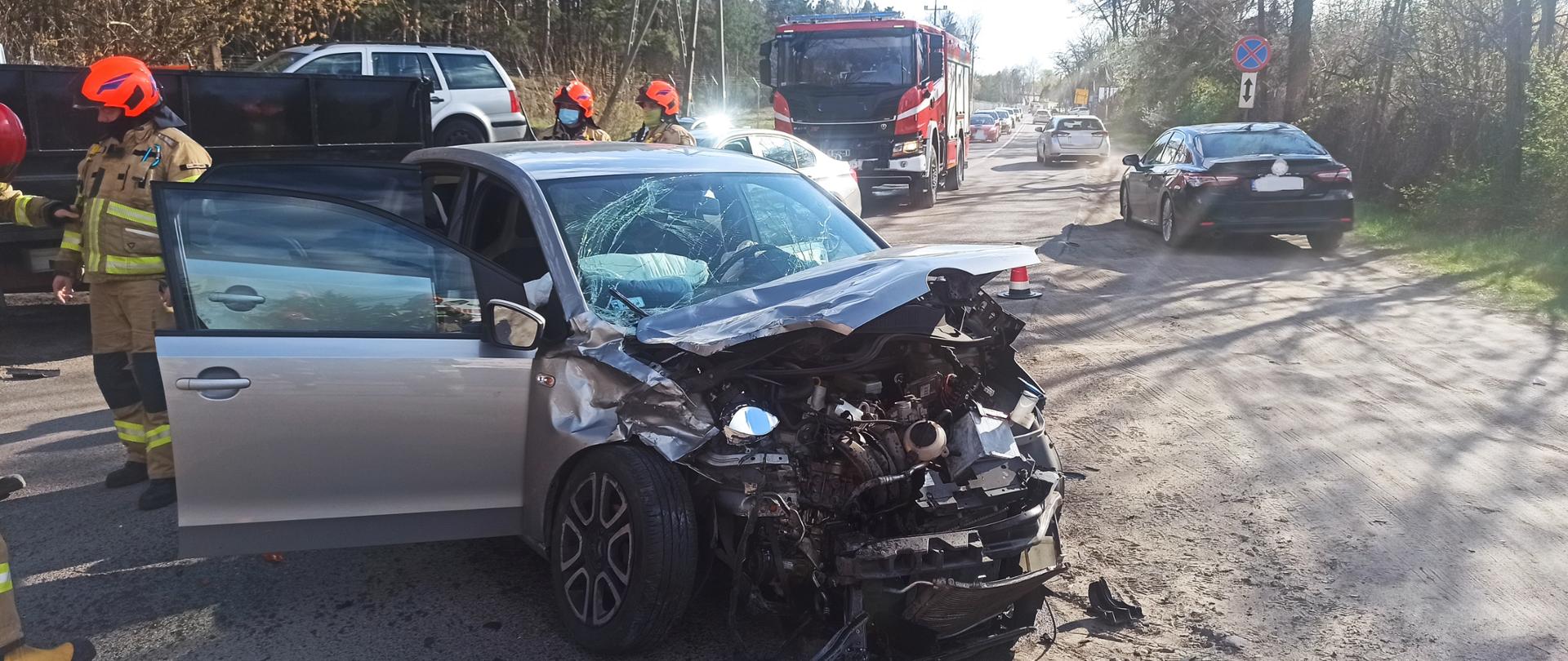 Na środku zdjęcia widać rozbity samochód osobowy koloru srebnego (uszkodzony przód i maska, drzwi po lewej stronie są otwarte na pełną szerokość). Pojazd jest na środku drogi. Obok pojazdu znajduje się trzech strażaków - w ubraniach specjalnych koloru piaskowego, na głowach noszą pomarańczowe hełmy . W głębi zdjęcia stoi samochód pożarniczy, a za nim kolumna samochodów osobowych. Na poboczu drogi są dwa samochody osobowe jeden srebrny, drugi szary. Po obu stronach drogi rosną drzewa.