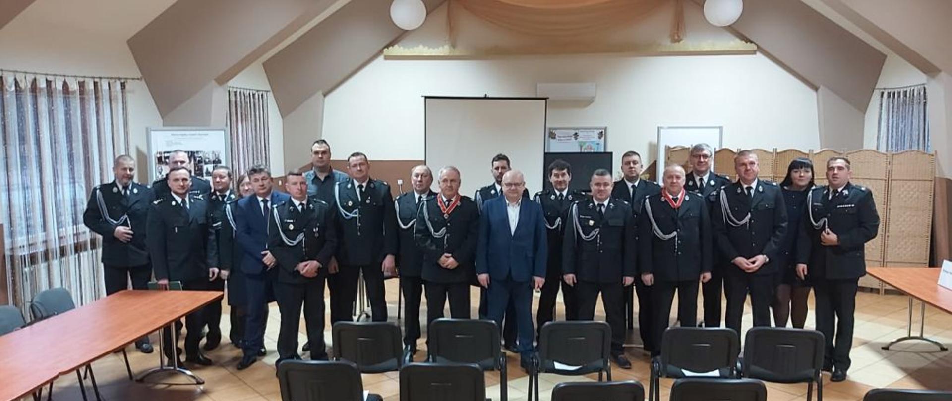 W pomieszczeniu na środku sali w dwuszeregu stoi kilkadziesiąt dh OSP w ciemnych mundurach. Przed nimi poustawiane krzesła po bokach soją stoły.