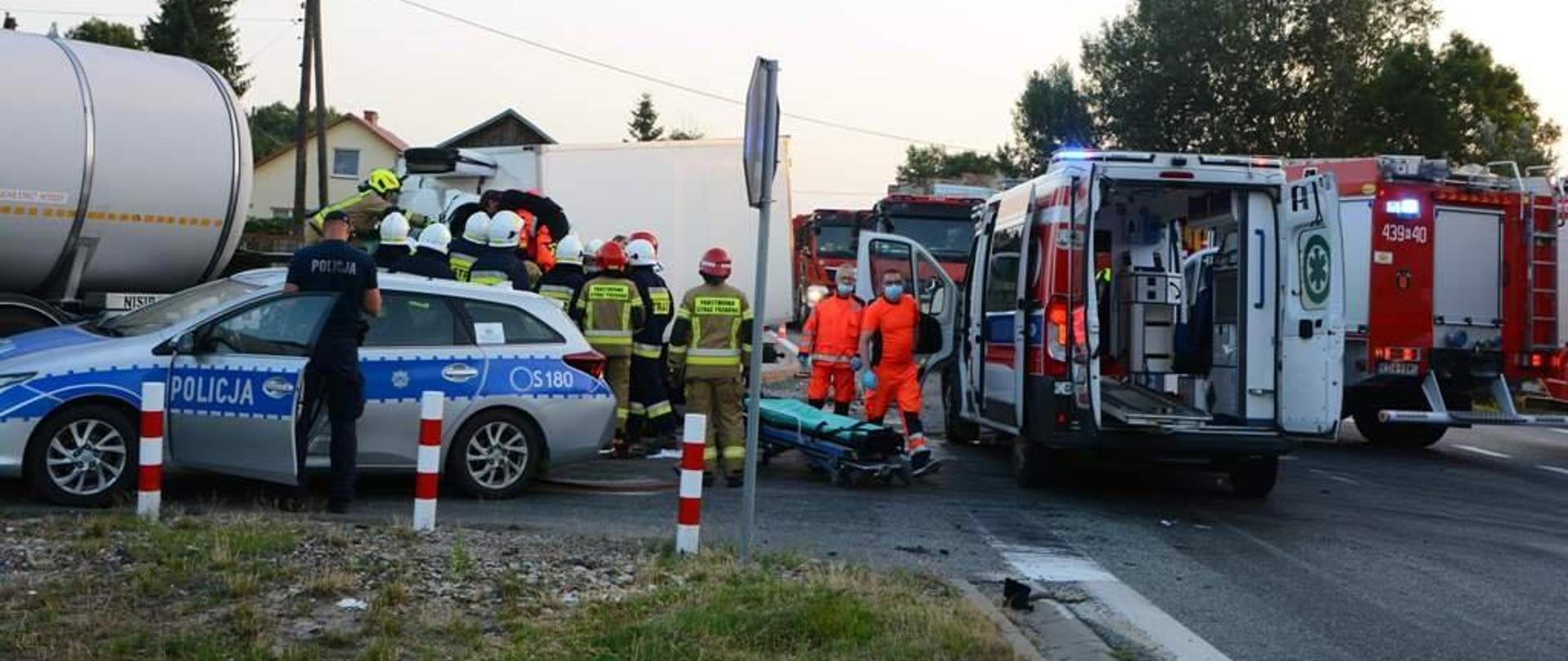 Na zdjęciu karetka pogotowia ratunkowego obok której dwóch ratowników medycznych przygotowuje nosze, radiowóz policyjny przy którym znajduje się policjant oraz grupa strażaków prowadząca działania ratownicze przy samochodzie dostawczym
