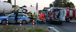 Na zdjęciu karetka pogotowia ratunkowego obok której dwóch ratowników medycznych przygotowuje nosze, radiowóz policyjny przy którym znajduje się policjant oraz grupa strażaków prowadząca działania ratownicze przy samochodzie dostawczym