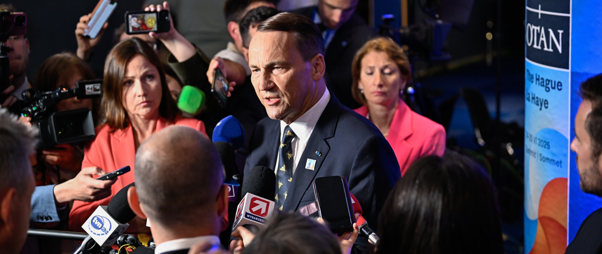 Mężczyzna w garniturze przemawia do kilku mikrofonów, w tle widoczny jest baner wydarzenia