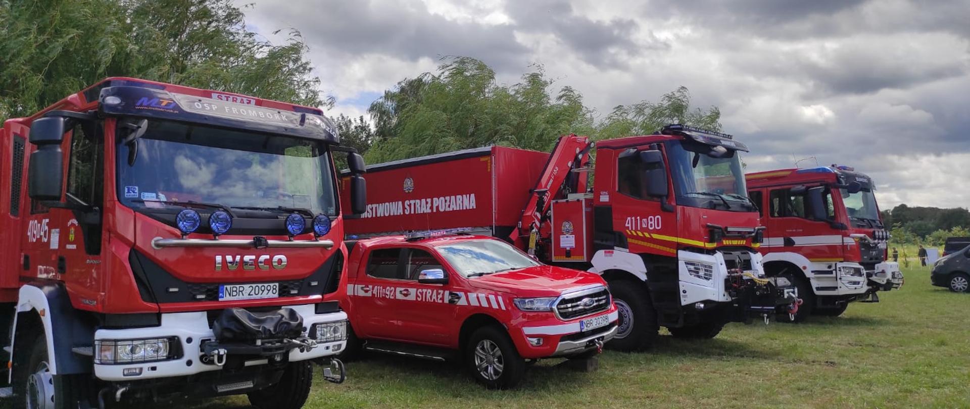 Na zdjęciu ustawione na polanie trawy w szeregu cztery pojazdy pożarnicze. Pochmurno.