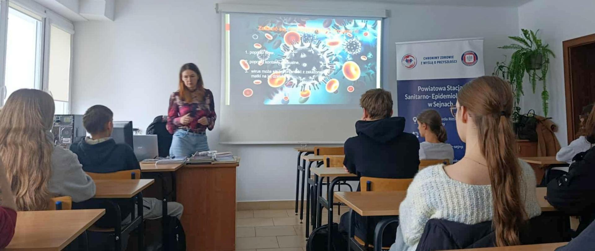 Sala lekcyjna, w której młoda kobieta prowadzi prezentację dla siedzącej publiczności, a na ekranie projektora wyświetlany jest slajd z grafiką wirusa i tekstem na temat zdrowia.