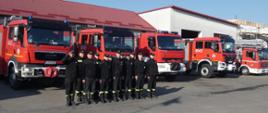 Na zdjęciu widać strażaków stojących na baczność w dwuszeregu, ubranych w ubranie koszarowe. Dowódca pododdziału oddaje honor salutując. Za strażakami widoczne są pojazdy gaśnicze.