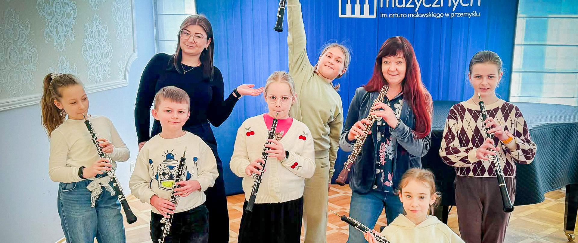 Grupa dzieci wraz z dwiema nauczycielkami stoi w jasnej sali muzycznej i trzyma oboje (instrumenty dęte drewniane). Jedna z dziewczynek unosi instrument wysoko do góry, inne dzieci trzymają swoje instrumenty w pozycji do gry. W tle widoczny jest niebieski baner szkoły muzycznej oraz fortepian. Wszyscy są ubrani na co dzień i wyglądają na zadowolonych.