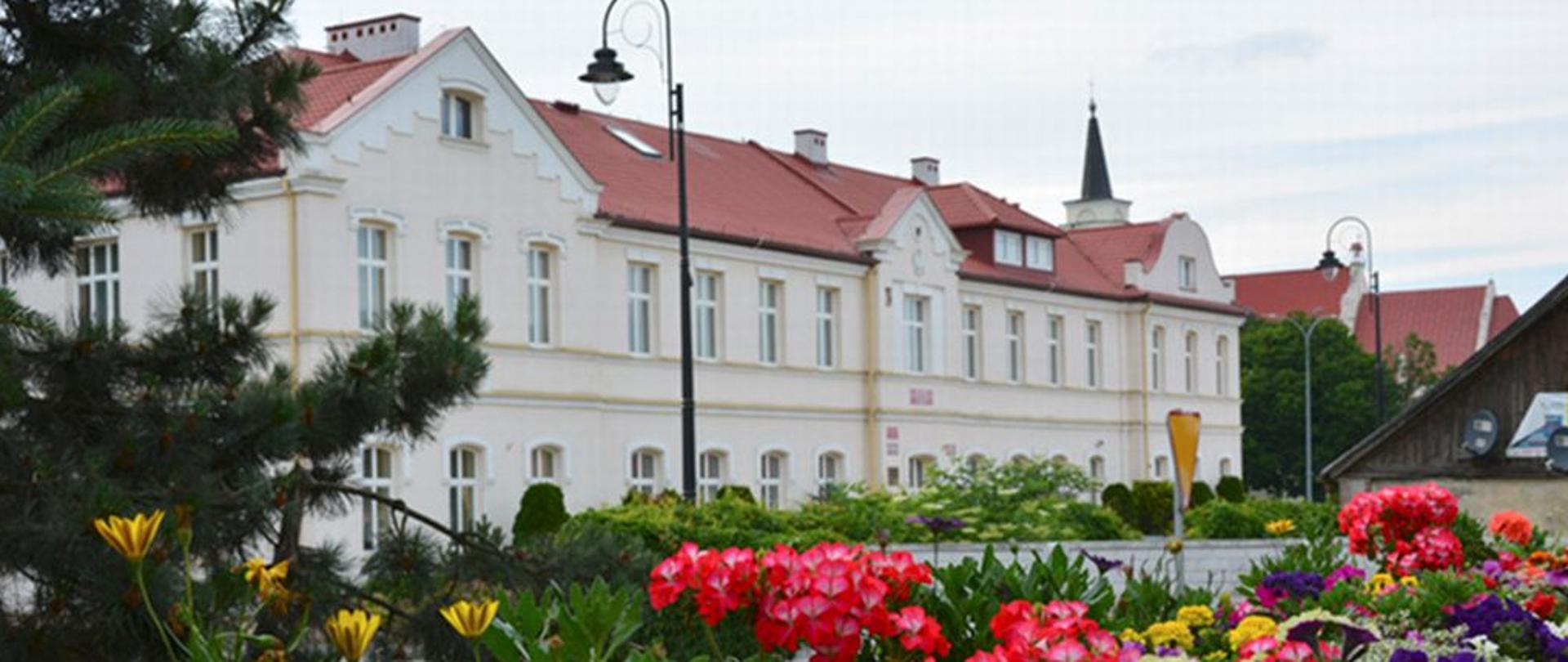 Budynek szkoły. Po lewej stronie choinka. Na dole zdjęcia kwiaty.