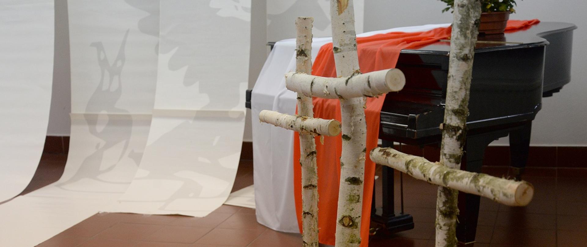 zdjęcie przedstawiające trzy krzyże z brzozy na tle fortepianu i leżącej na nim biało czerwonej flagi 