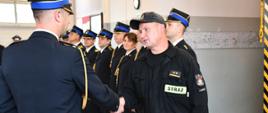 Na zdjęciu strażak w czarnym ubraniu koszarowym podaje rękę strażakowi mundurze w tle inni strażacy