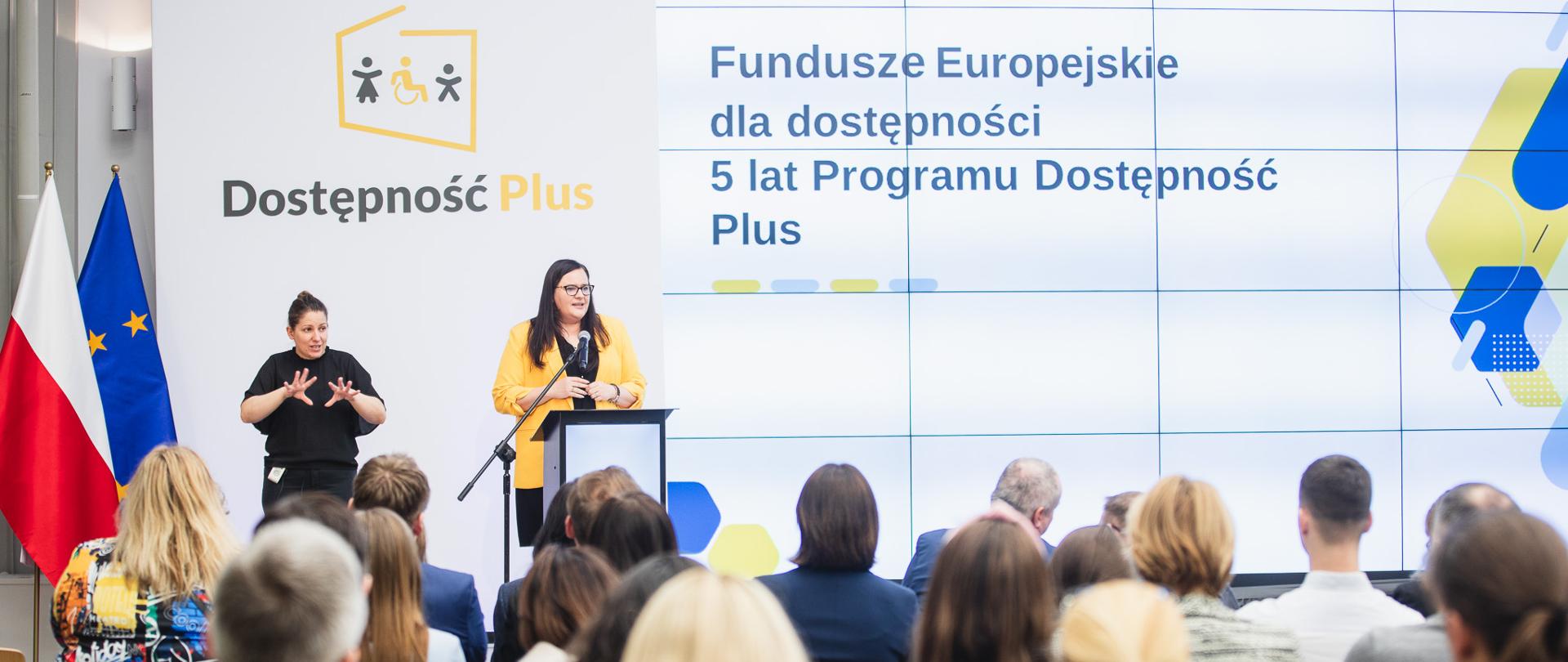 W sali konferencyjnej wiceminister Małgorzata Jarosińska-Jedynak w mównicy na tle ścianki z napisem Dostępność Plus. Obok tłumacz migowy. Przed nimi publiczność.