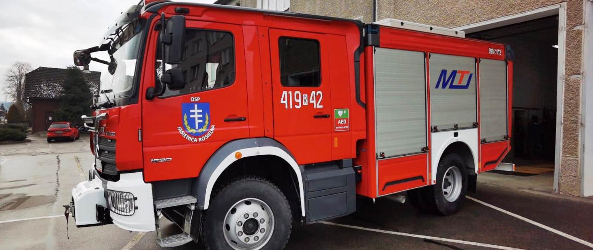 Kolorowe zdjęcie przedstawia na pierwszym planie nowy średni pojazd ratowniczy-gaśniczy GBA w OSP w Jasienicy Rosielnej. Samochód ciężarowy stoi na asfaltowym placu manewrowym przed brama garażową budynku Ochotniczej Straży Pożarnej w Jasienicy Rosielnej. W tle szara ściana budynku remizy z oknami i otwarta bramą garażową. W środkowej części fotografii widoczna częściowo przednia strona i lewa strona czerwonego samochodu ze srebrnymi żaluzjami zamkniętych skrytek na sprzęt w tylnej części zabudowy. Widoczny na nich niebiesko-czerwony napis MTI. Na środkowej części pojazdu biały numer operacyjny 419R42 a na drzwiach kierowcy biało-niebieski symbol i napis Jasienica Rosielna. 