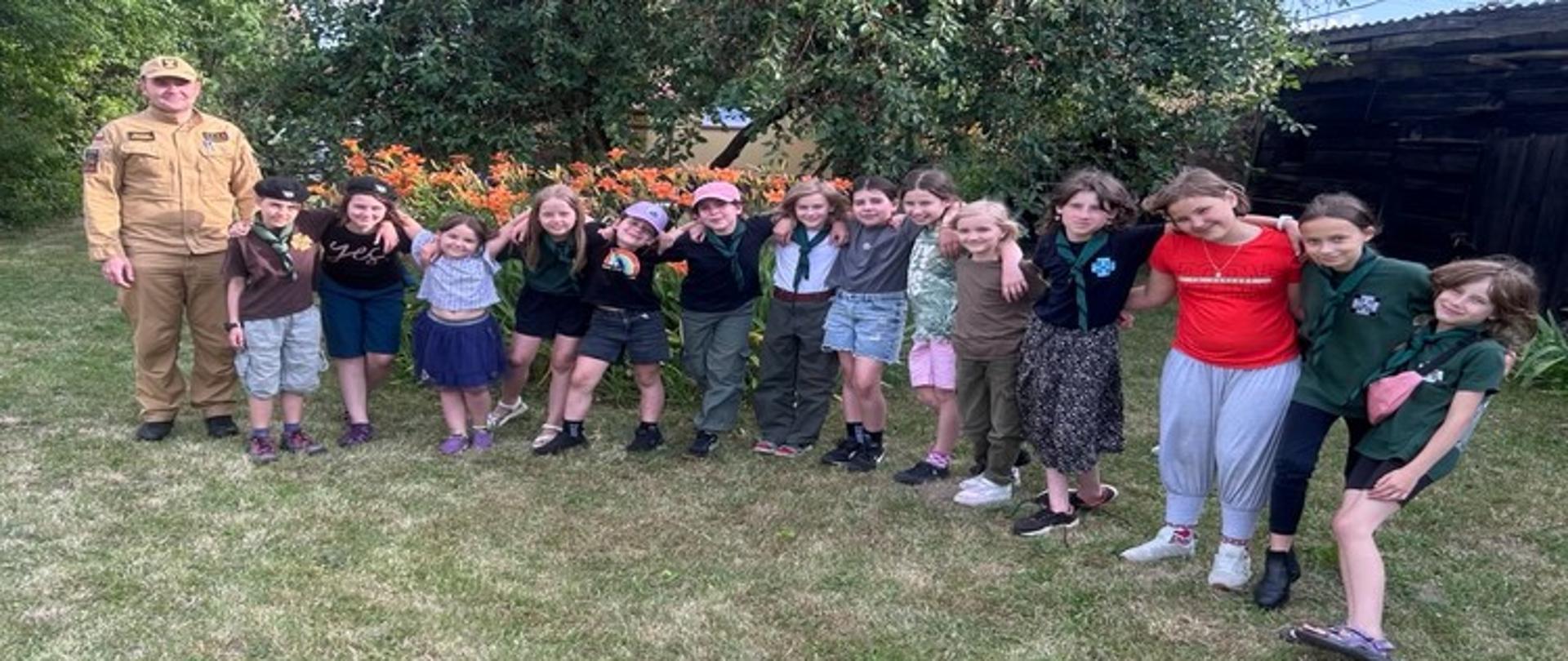 Zdjęcie grupowe strażaka z harcerkami. Na tle zielonej trawy, kwitnących kwiatów i drzew za nimi. Część harcerek ubrana w umundurowanie harcerskie, część w ubraniach cywilnych. Wszyscy na zdjęciu są w dobrym humorze - uśmiechnięci.