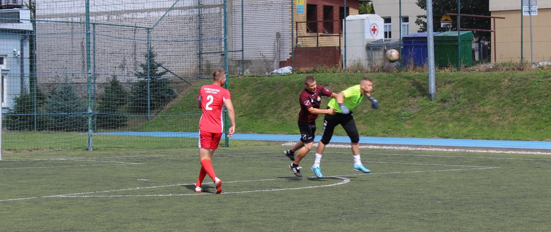 Na boisku piłkarskim znajduje się trzech piłkarzy. Bramkarz wybija piłkę za pomocą głowy. Przeciwnik próbuje mu w tym przeszkodzić.