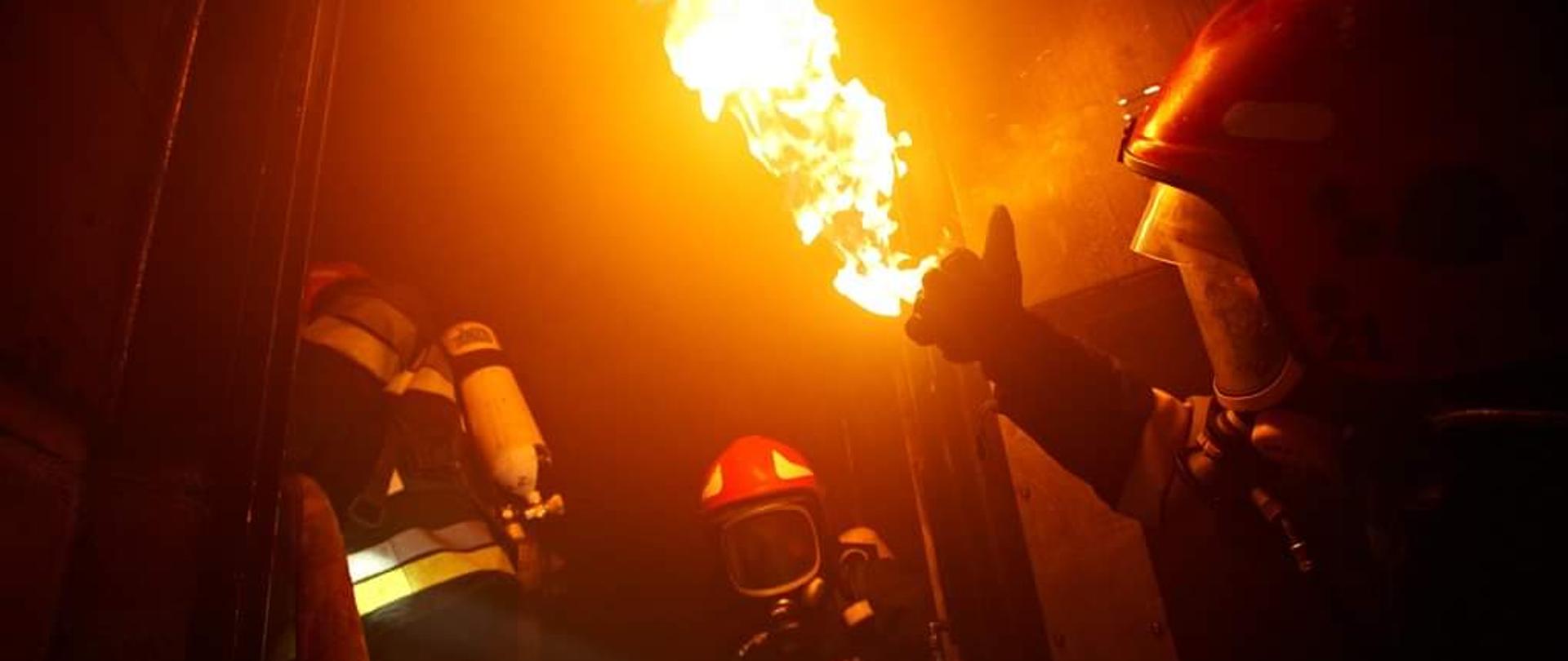  Ćwiczenia strażaków w komorze rozgorzeniowej, trzech strażaków w aparatach powietrznych oraz w hełmach robią unik przed płomieniem.