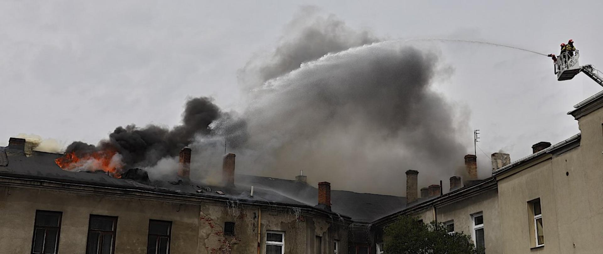 Zdjęcie przedstawia kamienicę oraz płomienie wydobywające się spod dachu. Obok strażacy na podnośniku podający wodę na pożar. Panuje duże zadymienie.