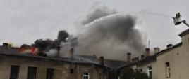 Zdjęcie przedstawia kamienicę oraz płomienie wydobywające się spod dachu. Obok strażacy na podnośniku podający wodę na pożar. Panuje duże zadymienie.