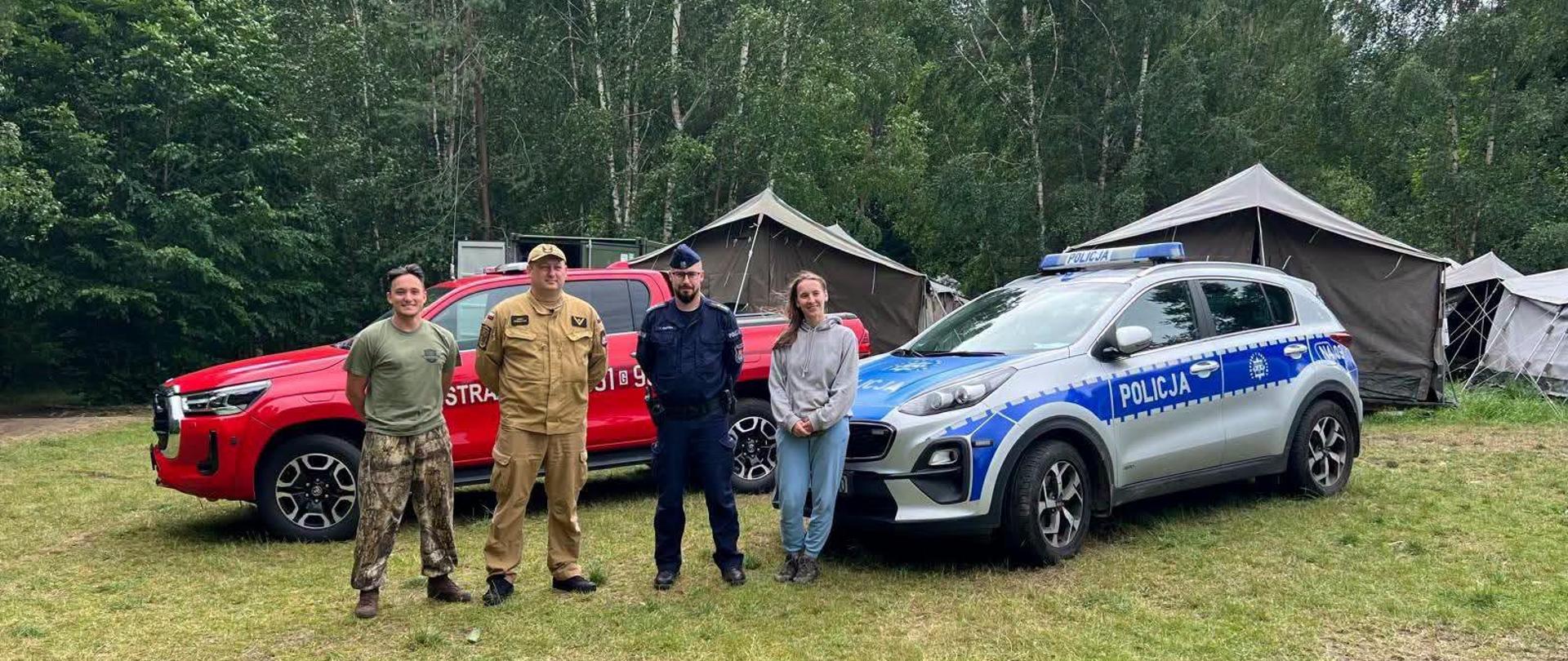 NA ZDJĘCIU WIDZIMY STRAŻAKA I POLICJANTA Z OPIEKUNAMI OBOZU HARCERSKIEGO NA TLE POJAZDÓW SŁUŻB