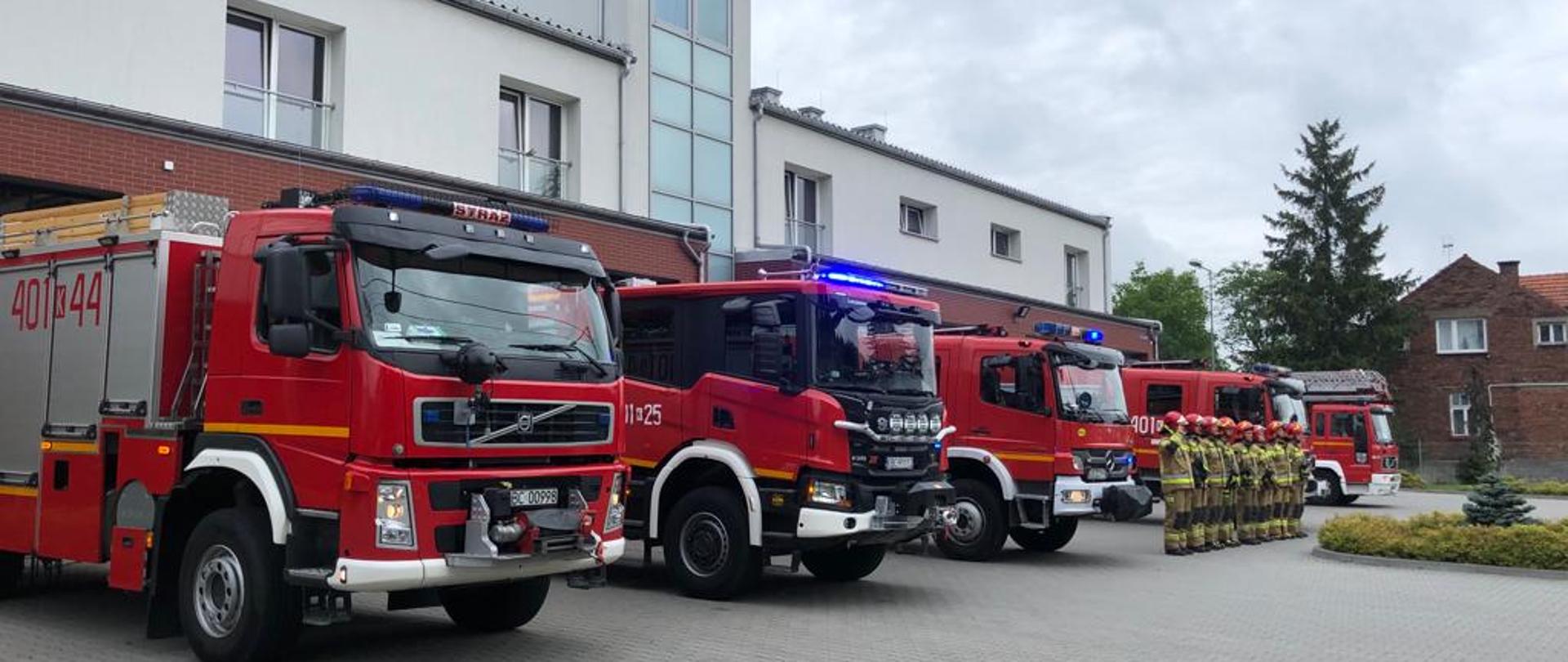 Zdjęcie przedstawia strażaków, którzy stoją na placu przed garażami z okazji zbiórki zorganizowanej z okazji tragicznej śmierci strażaka. Strażacy ubrani są w stroje bojowe czyli hełmu czerwone na głowie, ubrania specjalne koloru żółtego. Strażacy mają wzrok skierowany przed siebie. Z tyłu ustawione są samochody pożarnicze koloru czerwone go. W tle budynek komendy powiatowej