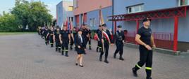 Strażacy trzymając poczty sztandarowe maszerują trójkami.
