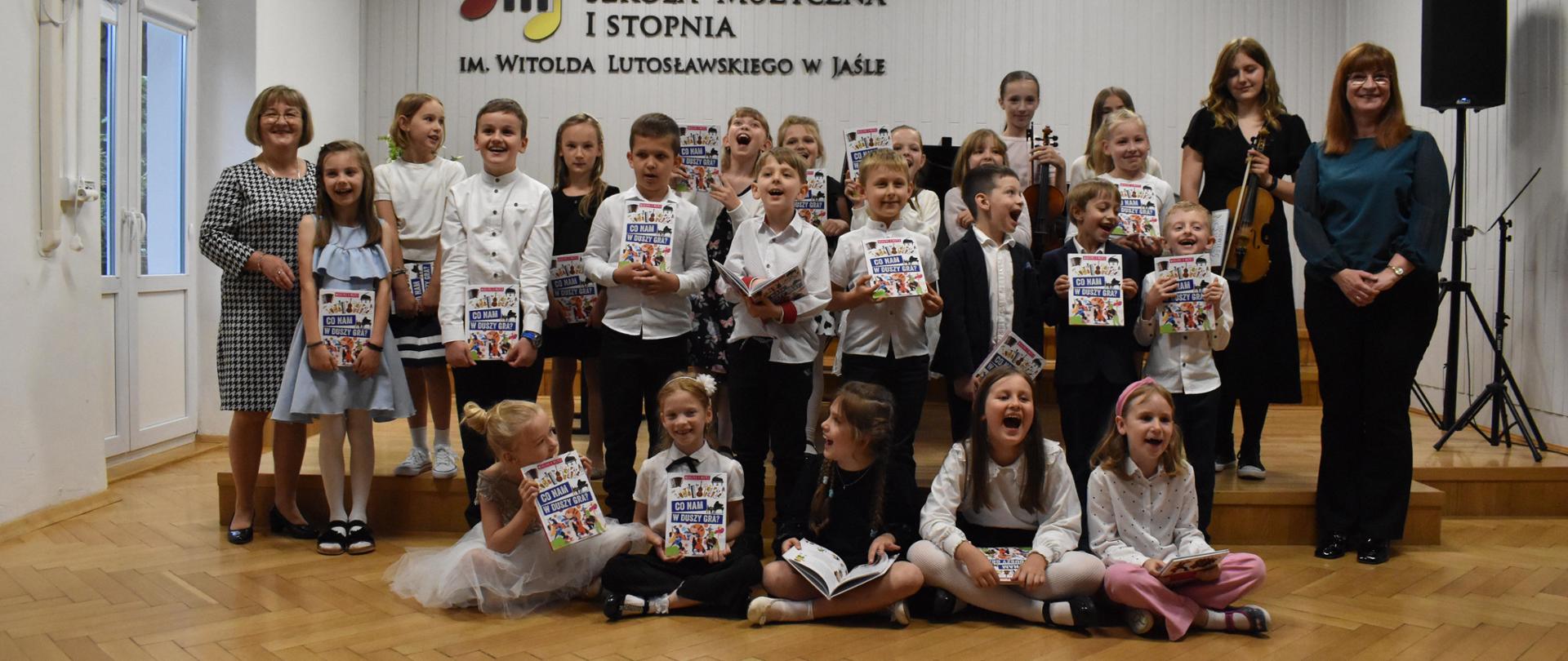 Na zdjęciu 23 dzieci, które trzymają w ręku książeczki oraz 2 osoby dorosłe. Wszyscy są uśmiechnięci. W tle zdjęcia napis "Państwowa Szkoła Muzyczna Im. Witolda Lutosławskiego w Jaśle" oraz godło Polski. Kolorystyka zdjęcia: na górze w odcieniach bieli, na dole w brązie, natomiast dzieci ubrane są na galowo.
