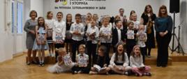 Na zdjęciu 23 dzieci, które trzymają w ręku książeczki oraz 2 osoby dorosłe. Wszyscy są uśmiechnięci. W tle zdjęcia napis "Państwowa Szkoła Muzyczna Im. Witolda Lutosławskiego w Jaśle" oraz godło Polski. Kolorystyka zdjęcia: na górze w odcieniach bieli, na dole w brązie, natomiast dzieci ubrane są na galowo.