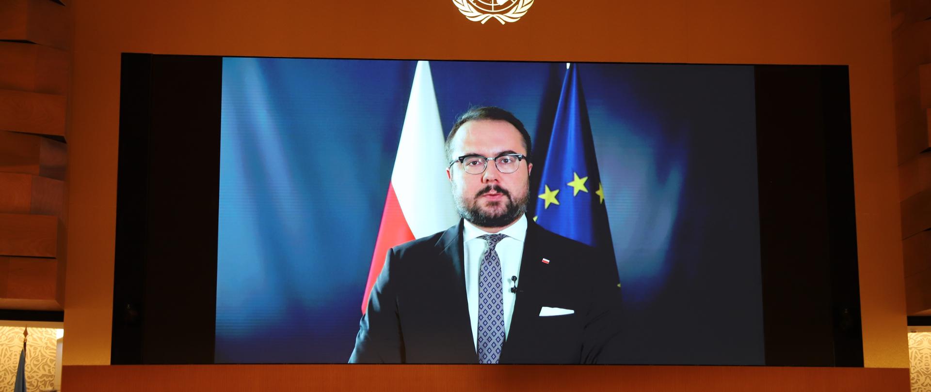 Duży monitor na ścianie z wyświetlanym obrazem brodatego mężczyzny w garniturze i krawacie na tle flagi polskiej i unijnej. Pod monitorem przy stole siedzą ludzie. 
