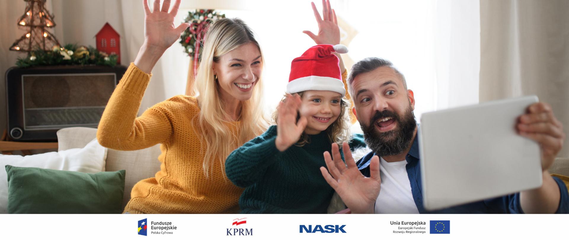 Trzyosobowa rodzina, dziecko w czapce św. Mikołaja siedzi na kanapie i macha do bliskich, z którymi połączyli się online. Mężczyzna trzyma w ręku tablet.