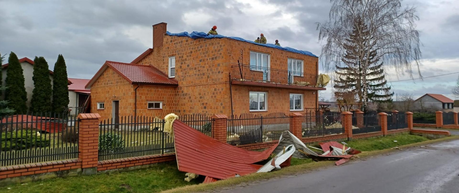 Na zdjęciu widać piętrowy dom z czerwonej cegły bez dachu. Przed domem w ogródku oraz w rowie leżą elementy dachu, blacha, wełna. Na dachu czterech strażaków w żółtych strojach i czerwonych hełmach rozkładają niebieską folię. W tle widać inne budynki oraz drzewa. Jest widno.