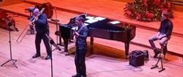 Na estradzie sali koncertowej stoi dwóch mężczyzna, jeden gra na puzonie, drugi na gitarze basowej. Za nimi stoi fortepian, obok po prawej stronie na wysokim krześle siedzi mężczyzna.