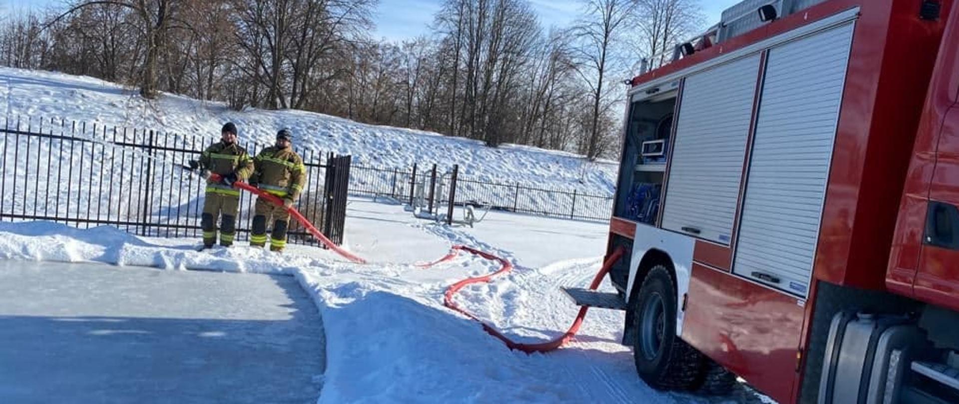 Dwóch funkcjonariuszy PSP w ubraniach specjalnych podają jeden prąd wody z samochodu gaśniczego na boisko sportowe, na którym jest utworzone sztuczne lodowisko. 