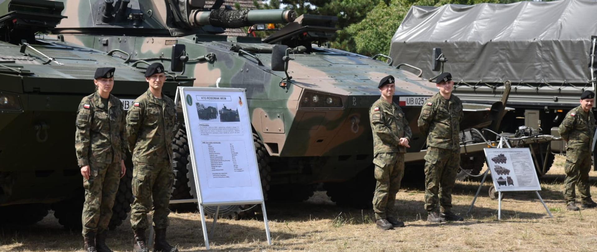 Na trawniku stoją pojazdy wojskowe. Przy każdym dwoje żołnierzy i tablica informacyjna