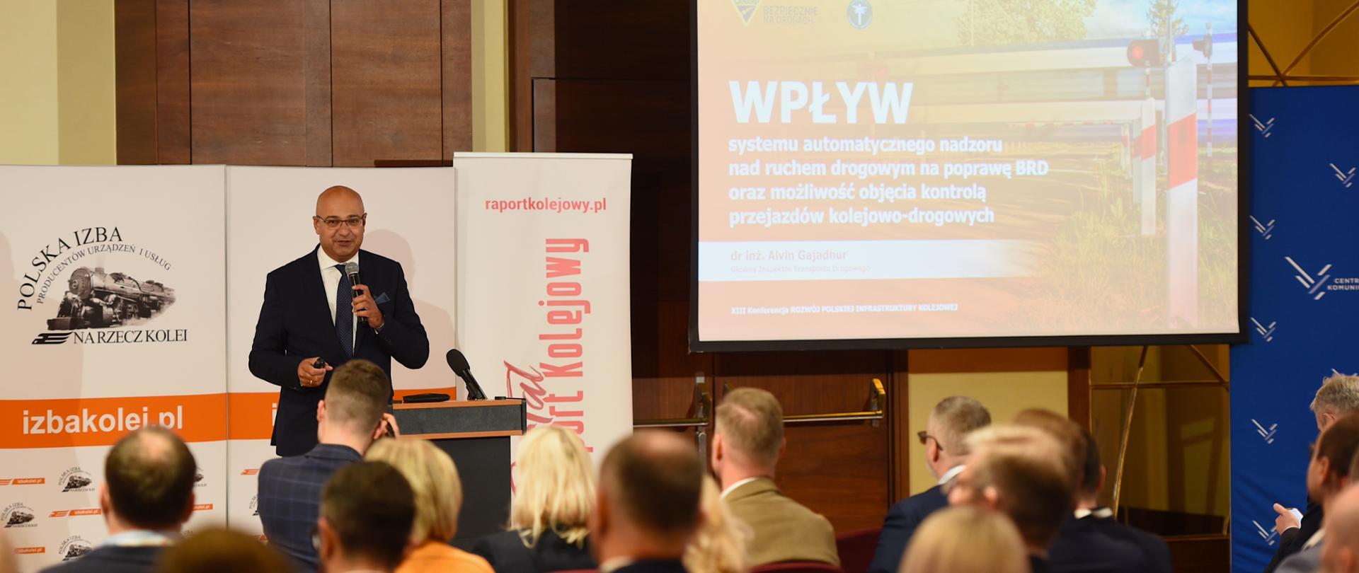 Alvin Gajadhur przemawia. Obok niego na ekranie prezentacja. 