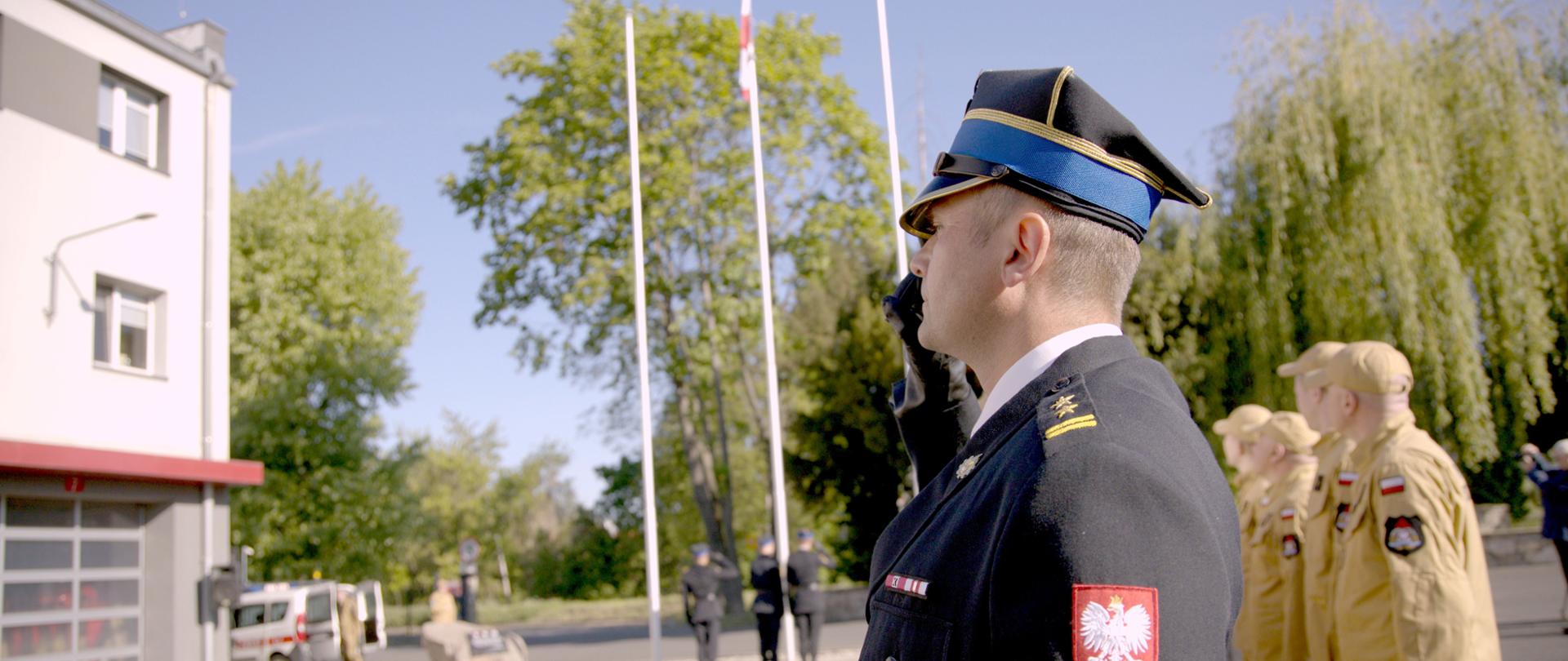 Na zdjęciu widać funkcjonariuszy w momencie podniesienia flagi na maszt. Na pierwszym planie dowódca uroczystości. Wszyscy funkcjonariusze w umundurowaniu służbowym. Uroczystość na placu zewnętrznym KW PSP w Opolu.