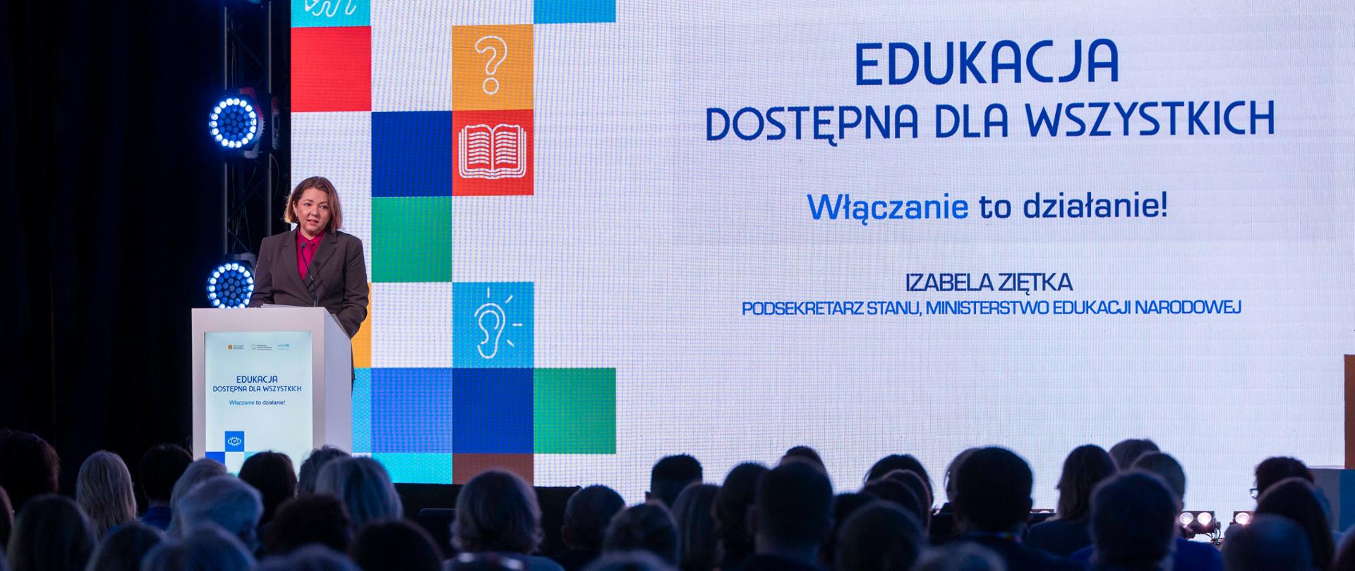 Kobieta (Podsekretarz Stanu Izabela Ziętka) w szarym garniturze stoi na mównicy i przemawia, z tytułem "EDUKACJA DOSTĘPNA DLA WSZYSTKICH. Włączanie to działanie!" oraz informacją o prelegentce. Scenę wypełniają duże, podświetlone kolorowe kwadraty z ikonami. Na pierwszym planie widoczna jest publiczność.