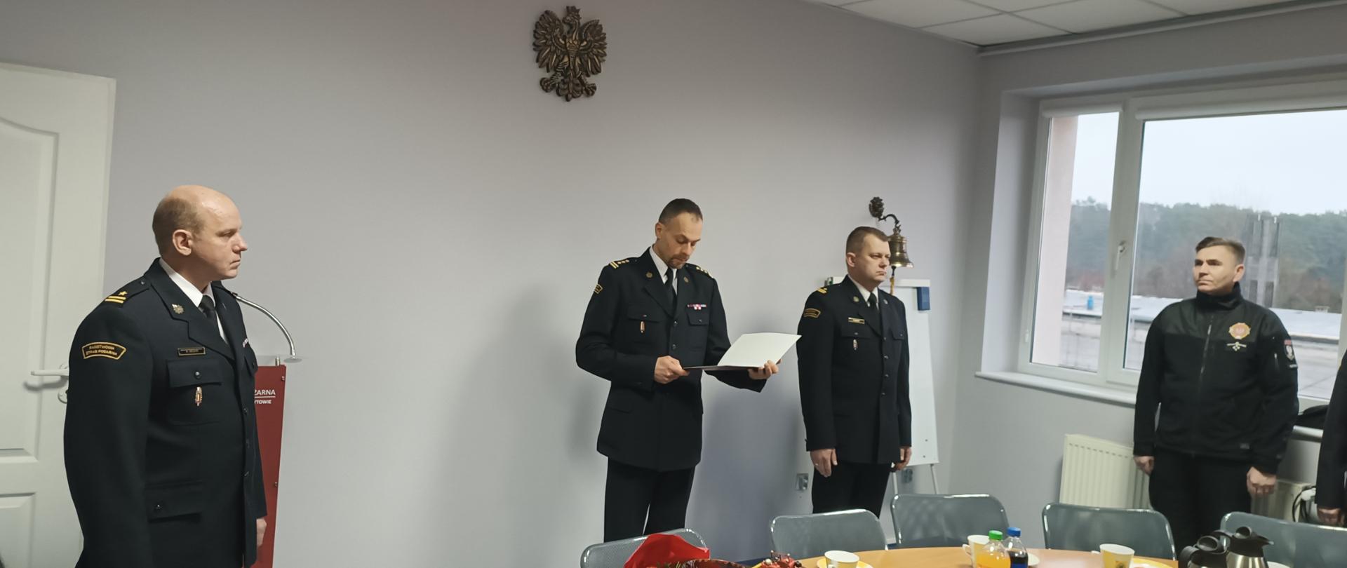 Czerech mężczyzn w ciemnych mundurach strażackich. Jeden z nich trzyma w ręku dokument, który odczytuje. 