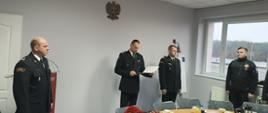 Czerech mężczyzn w ciemnych mundurach strażackich. Jeden z nich trzyma w ręku dokument, który odczytuje. 