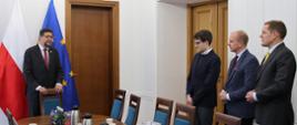 Wiceminister Szeptycki stoi przy podłużnym stole i mówi do trzech stojących obok mężczyzn.