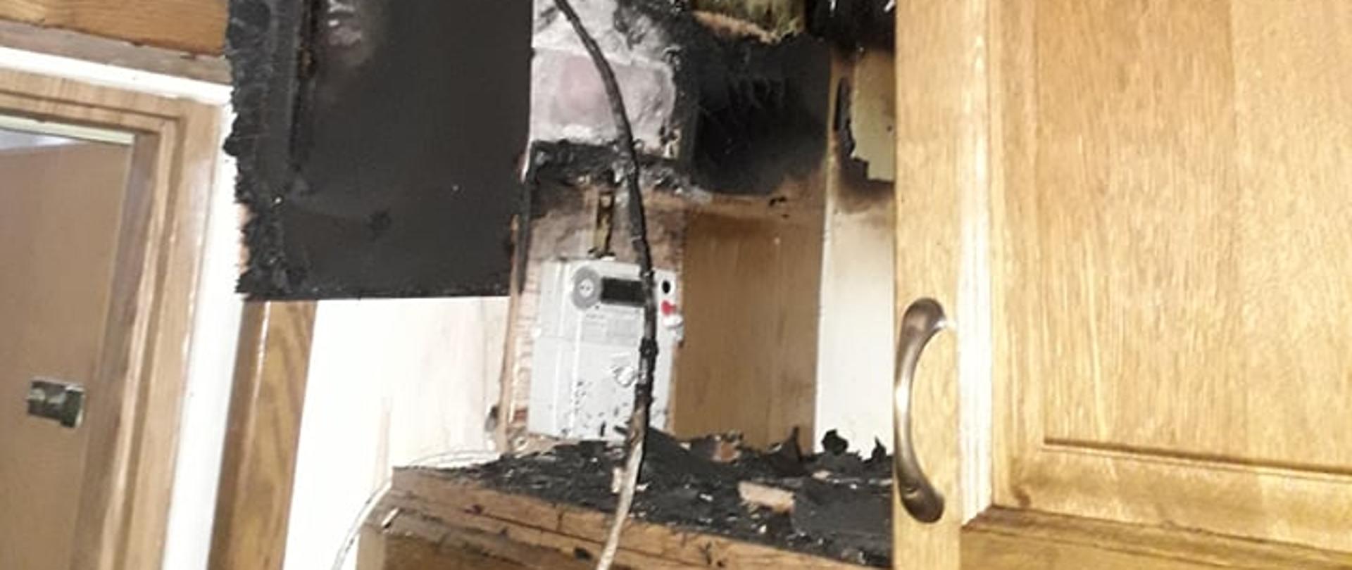 Na zdjęciu widoczne są szafki drewniane, których część uległa spaleniu. W szafce widoczne są przewody elektryczne oraz licznik energii elektrycznej koloru białego. W górnej części zdjęcia widoczny jest pokryty sadzą sufit zaś po lewej stronie otwarte drzwi wejściowe do mieszkania. 