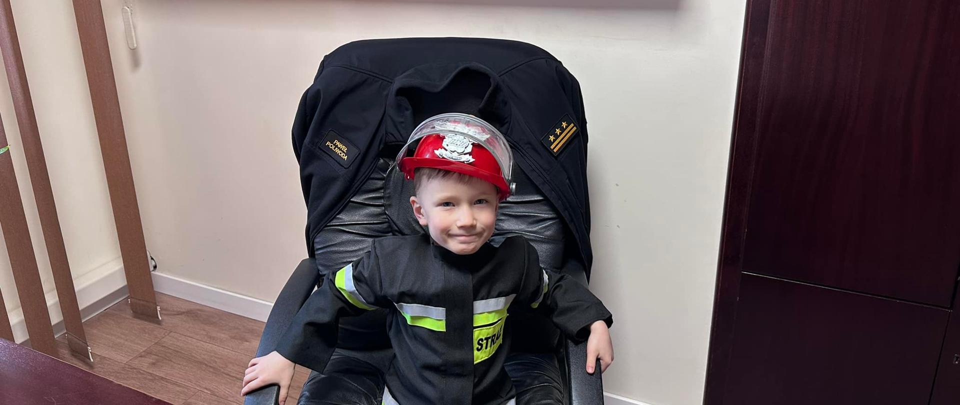 Na zdjęciu znajduje się chłopczyk ubrany w strój strażacki siedzący na fotelu przy biurku. W tle widać półkę z pucharami i zdjęciami