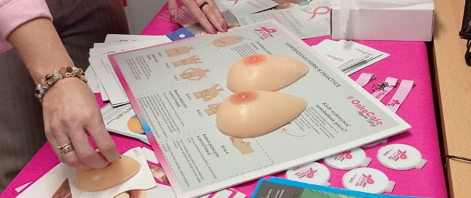 Zdjęcie przedstawia stół przykryty różowym obrusem. Na stole znajdują się materiały edukacyjne związane z badaniem raka piersi.
