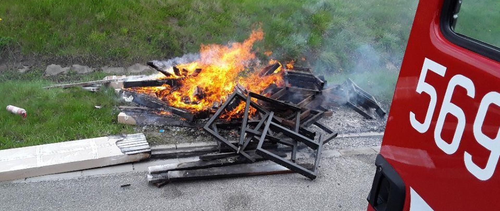 Na zdjęciu widoczne płonące materiały (stelaże, okopcone drewniane elemety) na poboczu drogi poza samochodem.