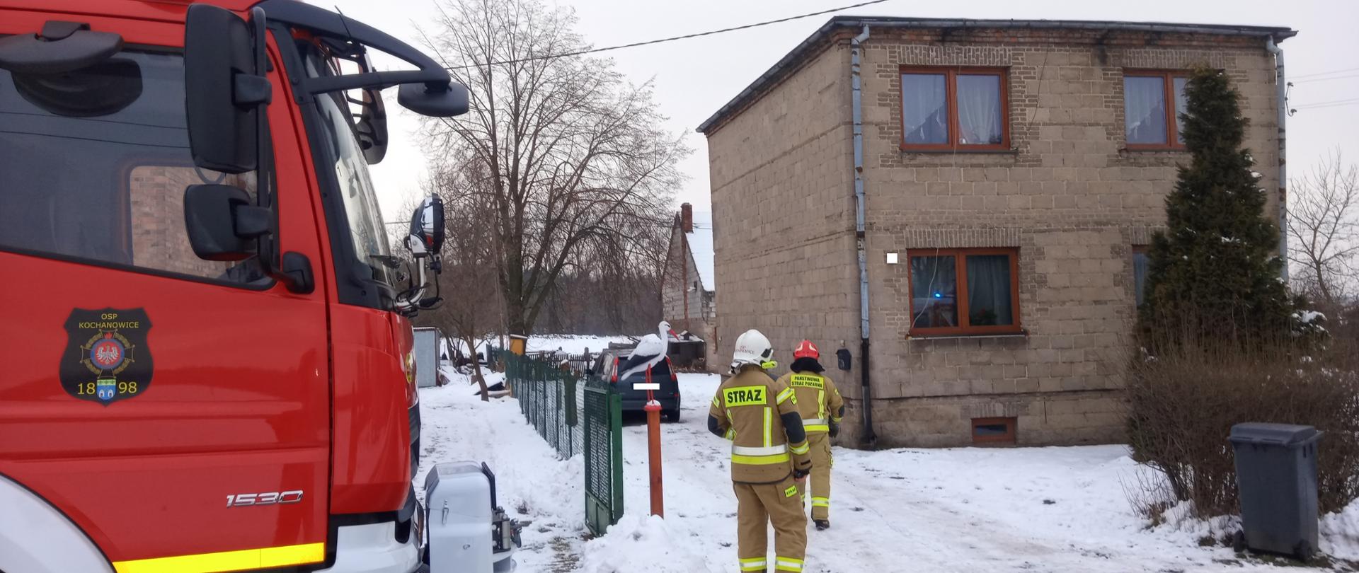 Zdjęcie zrobione na tle jednorodzinnego budynku mieszkalnego. Przed budynkiem dwóch strażaków w umundurowaniu specjalnym w hełmie. Po lewej stronie przód samochodu pożarniczego.