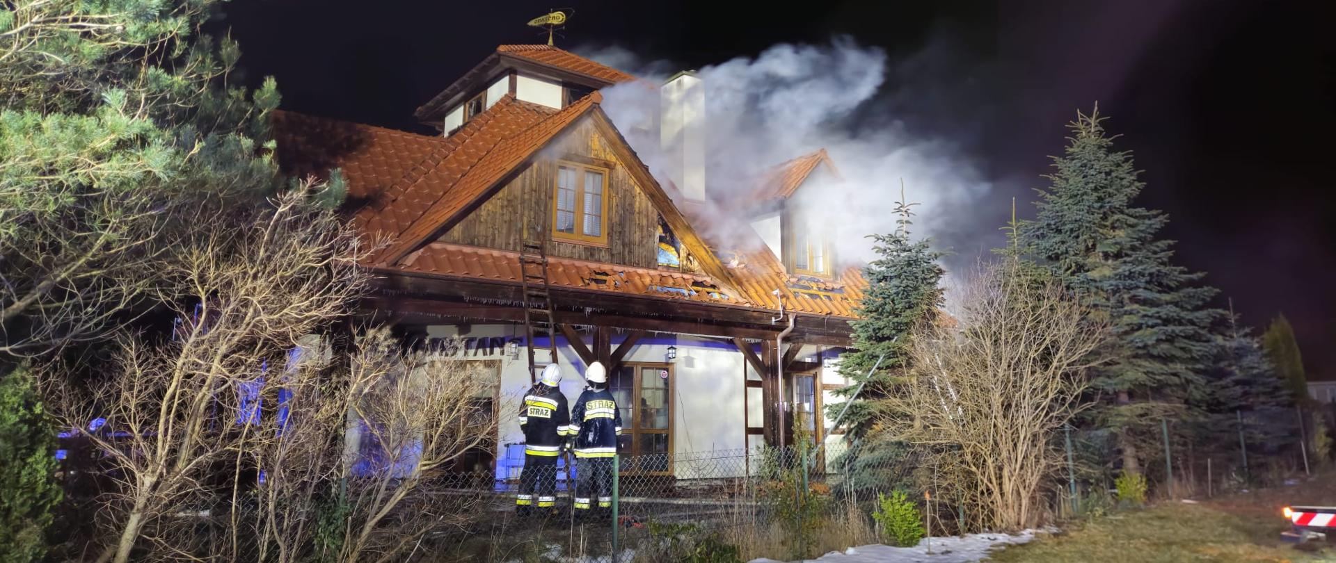 Strażacy w ubraniach specjalnych, ustawiają drabinę nasadkową przed budynkiem objętym pożarem, widoczne silne zadymienie