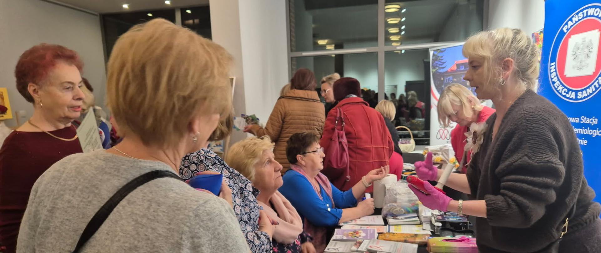 na zdjęciu kilka osób stoi wokół stolika, na którym rozłożone są ulotki dotyczące zdrowia. po prawej stronie na ścianie logo państwowej inspekcji sanitarnej