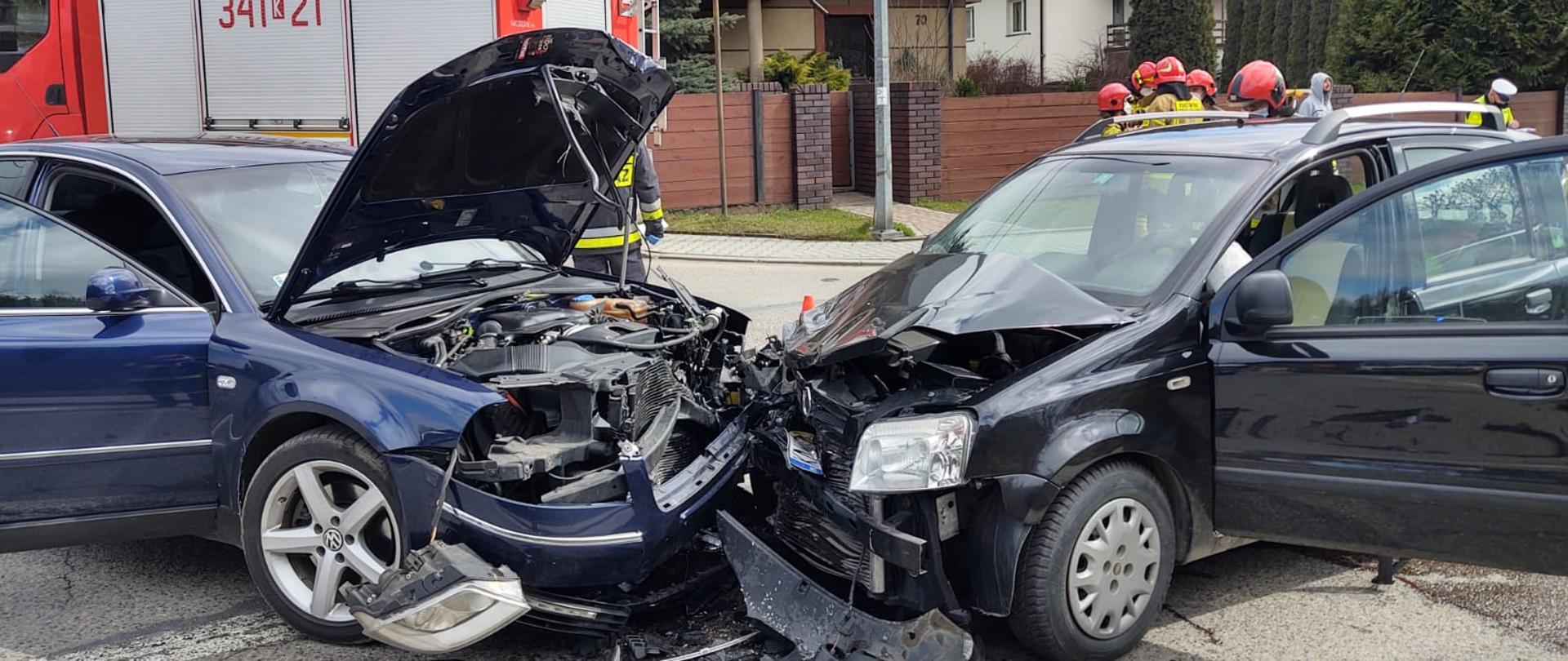 Widok dwóch samochodów, które uległy zderzeniu. W tle stoją strażacy oraz samochód pożarniczy. 