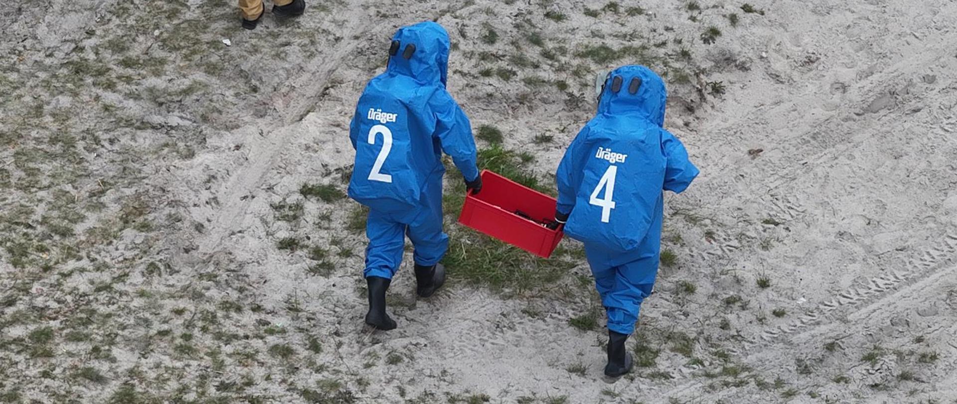 Dwóch strażaków w niebieskich skafandrach gazoszczelnych Dräger (z numerami 2 i 4) idzie przez piaszczysty teren, niosąc wspólnie czerwoną skrzynkę. Widok z drona.