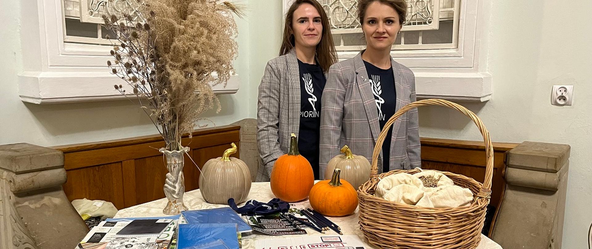 Dwie kobiety stojące za stołem, na którym znajdują się różne przedmioty. Kobiety mają na sobie ciemne koszulki z nadrukiem oraz marynarki w szarą kratkę. Na stole znajdują się dwie pomarańczowe dynie i jedna beżowa, obok nich kosz wiklinowy z materiałowymi woreczkami w środku. Po lewej stronie widzimy suchą kompozycję roślinną w wazonie. Na stole leżą także ulotki, broszury, długopisy i inne materiały informacyjne. Na froncie stołu znajduje się kartka z napisem „WOJEWÓDZKI INSPEKTORAT OCHRONY ROŚLIN I NASIENNICTWA W KIELCACH ODDZIAŁ W BUSKU-ZDROJU”. Całość odbywa się w jasnym, klasycznie urządzonym wnętrzu z ozdobnym oknem w tle.