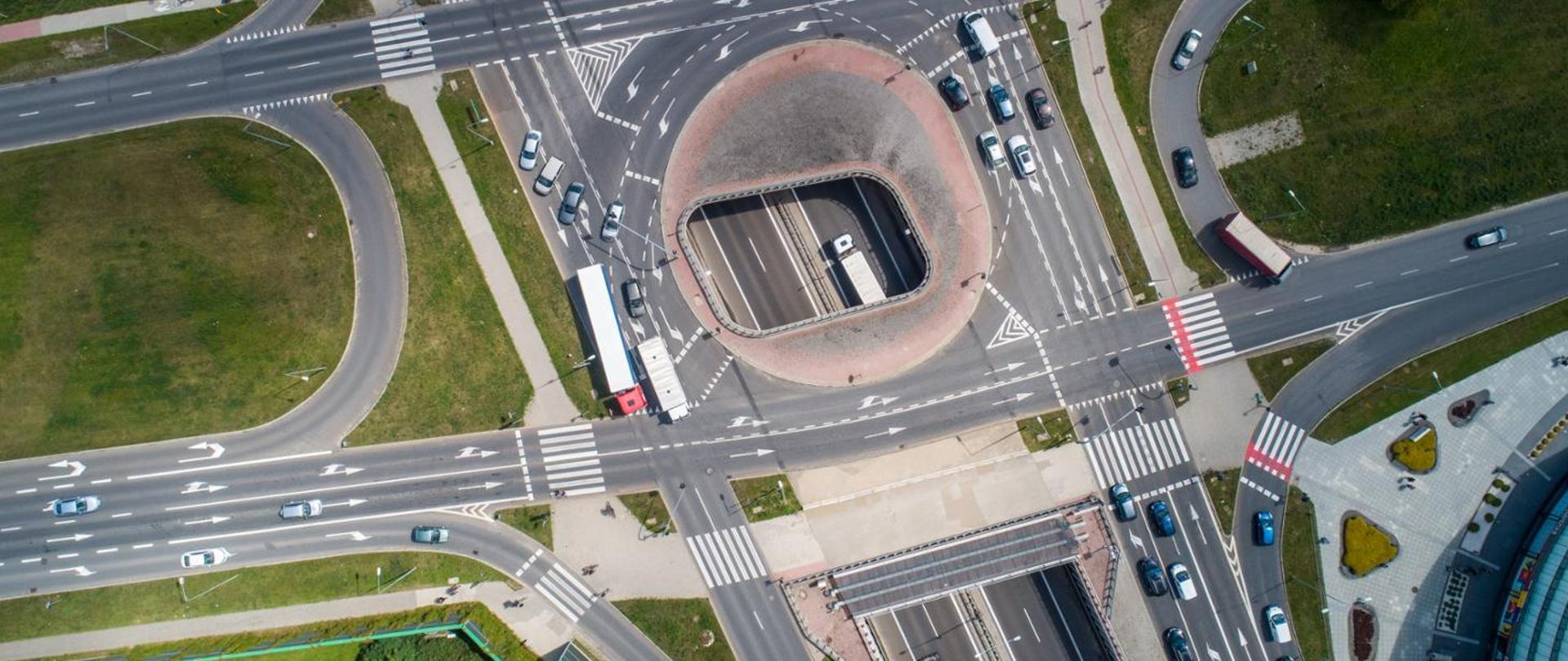 Widok z lotu ptaka na duże skrzyżowanie drogowe z rondem; wielopasmowe jezdnie z oznakowaniem poziomym, pasami do skrętu, przejściami dla pieszych i ścieżkami rowerowymi, po drogach poruszają się samochody, a wokół skrzyżowania widoczne są tereny zielone oraz chodniki.