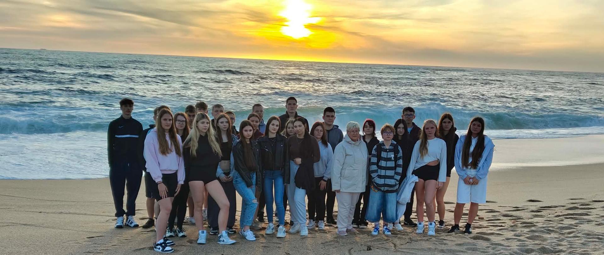 Grupa uczniów z nauczycielką pozuje do zdjęcia na tle zachodu słońca nad oceanem. Na piasku widnieje napis: ZSCKR Sejny.