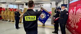 Uroczystość ślubowania strażaków w garażu Komendy Powiatowej Państwowej Straży Pożarnej w Obornikach. Funkcjonariusze w piaskowych ubraniach służbowych stoją w szeregu. Na tle czerwonego baneru PSP, odbywa się uroczyste ślubowanie.
